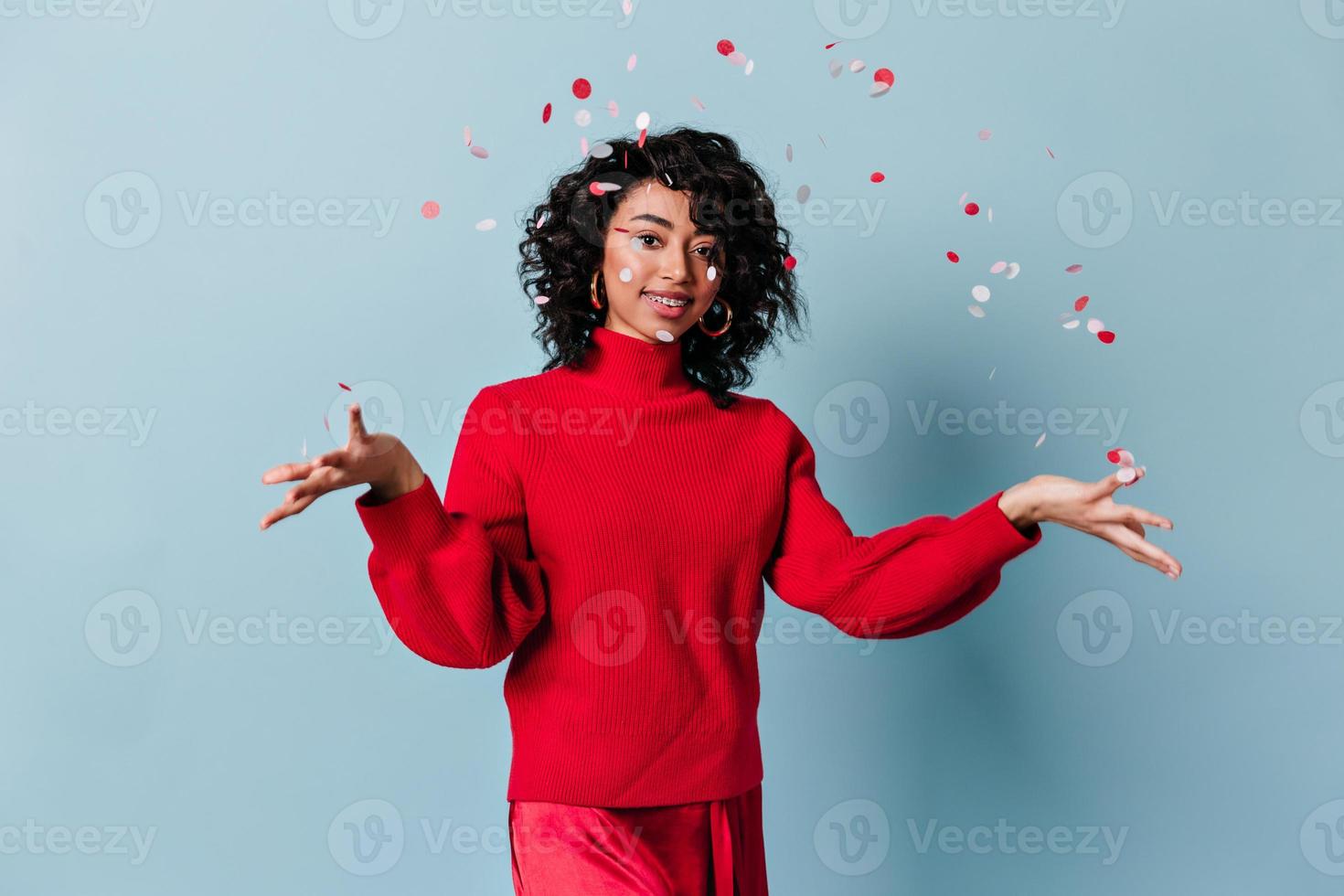 glückliches Mischlingsmädchen, das Konfetti auswirft. Studioaufnahme einer erstaunlichen Frau im roten Pullover. foto