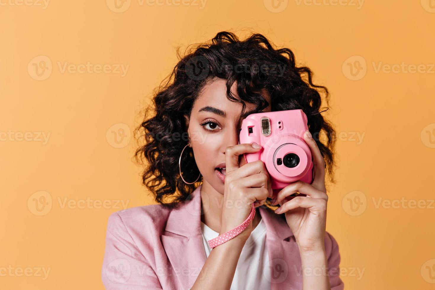 sinnliche Mischlingsfrau, die Fotos macht. Vorderansicht des glamourösen lockigen Mädchens mit rosa Kamera.
