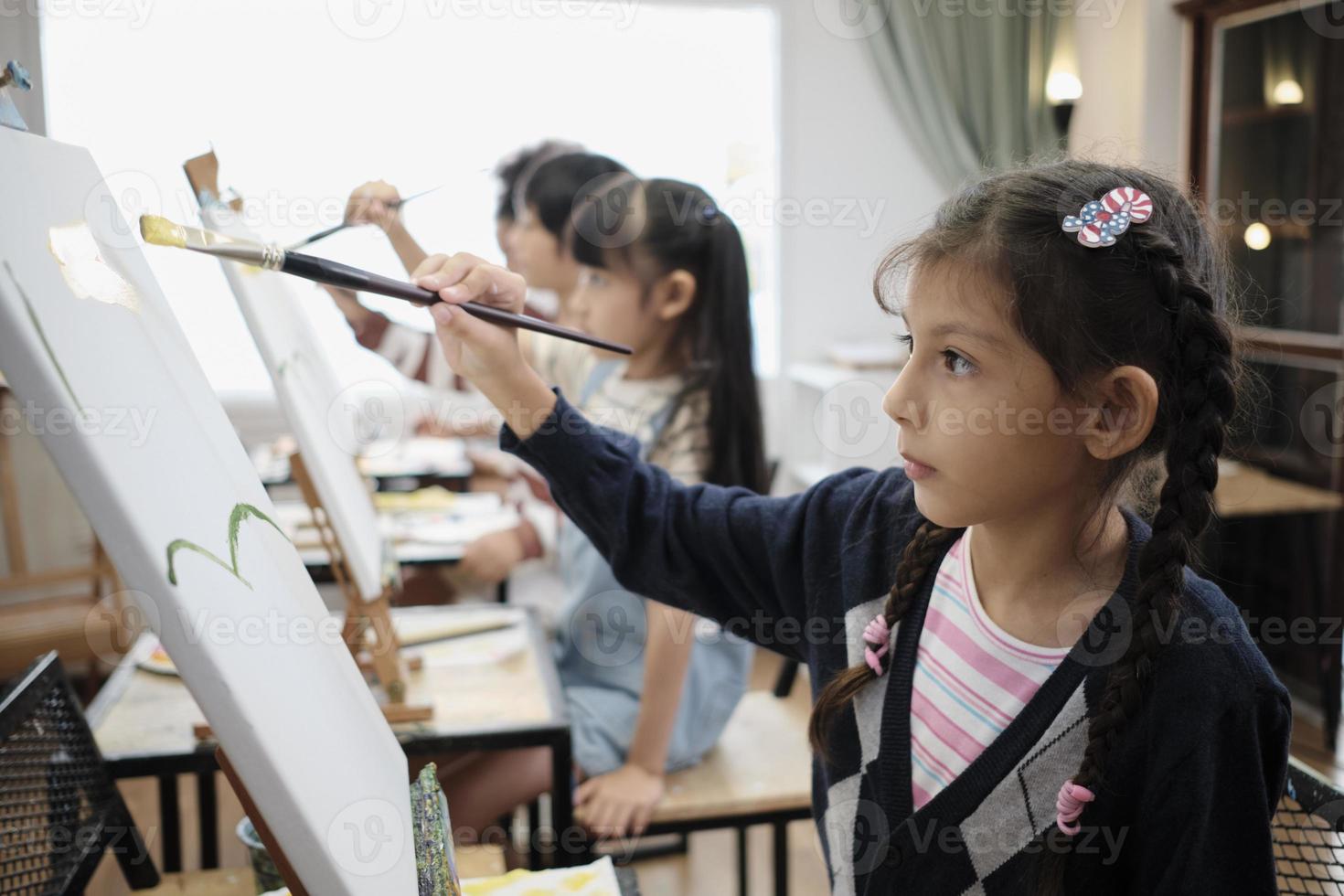 Ein kleines Mädchen konzentriert sich auf das Malen von Acrylbildern auf Leinwand mit gemischtrassigen Kindern in einem Kunstklassenzimmer, kreatives Lernen mit Talenten und Fähigkeiten in der Studioausbildung der Grundschule. foto