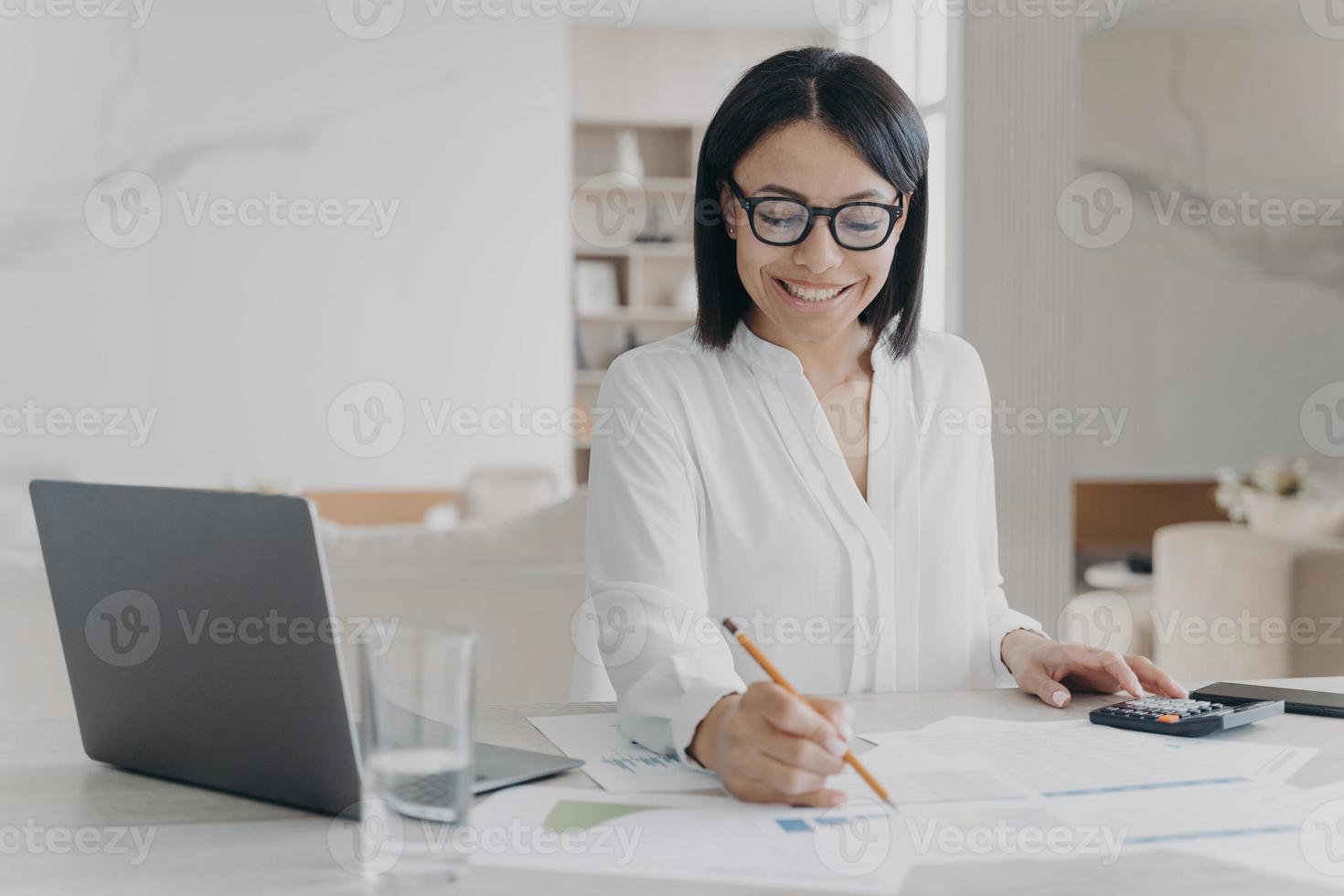 Lächelnde Geschäftsfrau mit Brille arbeitet bei der Berechnung des Projektbudgets und sitzt am Schreibtisch mit Laptop foto