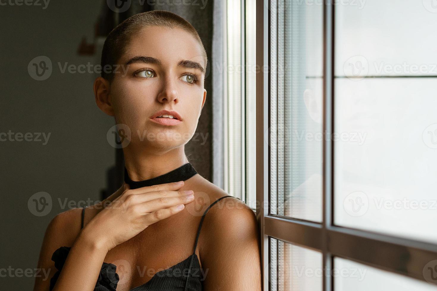 Porträt einer traurigen jungen Frau mit kurzen Haaren, die in einem schwarzen Kleid aus dem Fenster schaut foto
