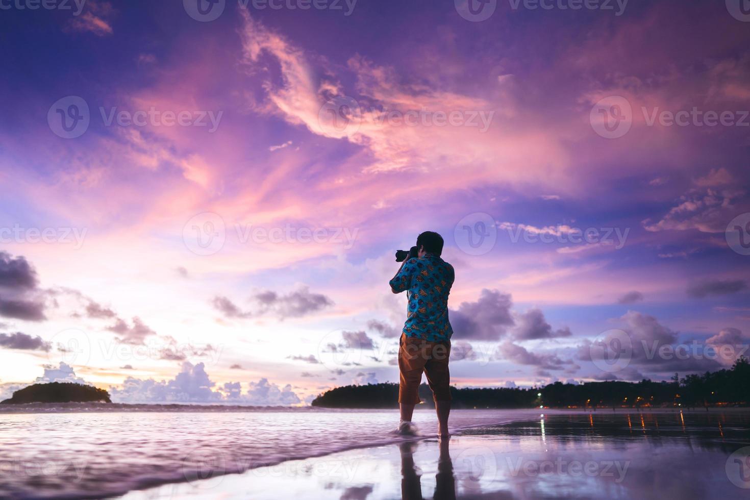 Rückansicht des asiatischen Fotografen für Erwachsene mit Kamera auf Strandsand mit wunderschönem dramatischem Sonnenuntergangshimmel foto