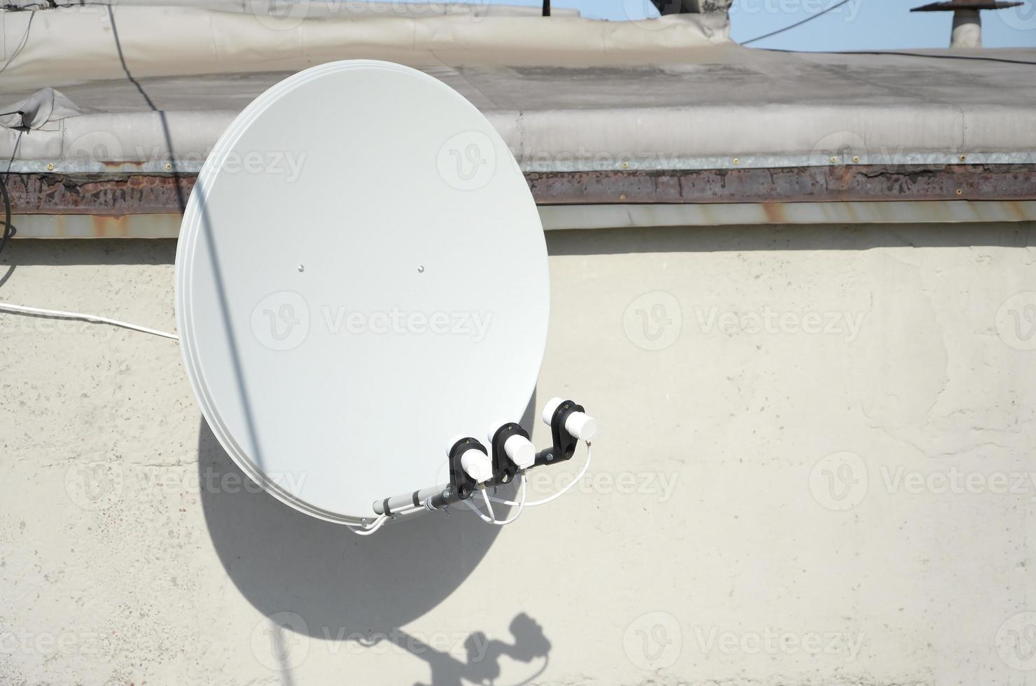 Weiße Satellitenschüssel mit drei Konvertern, die auf einer Betonwand auf dem Dach eines Wohngebäudes montiert sind. Satelliten Fernsehen foto