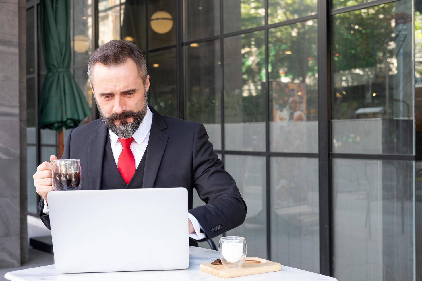 Außenaufnahme eines kaukasischen Geschäftsmannes, der heißen Morgenkaffee im Café trinkt, während er mit einem Computer-Laptop arbeitet und seine E-Mails überprüft. arbeitsroutine mit städtischem geschäftsgebietshintergrund. foto