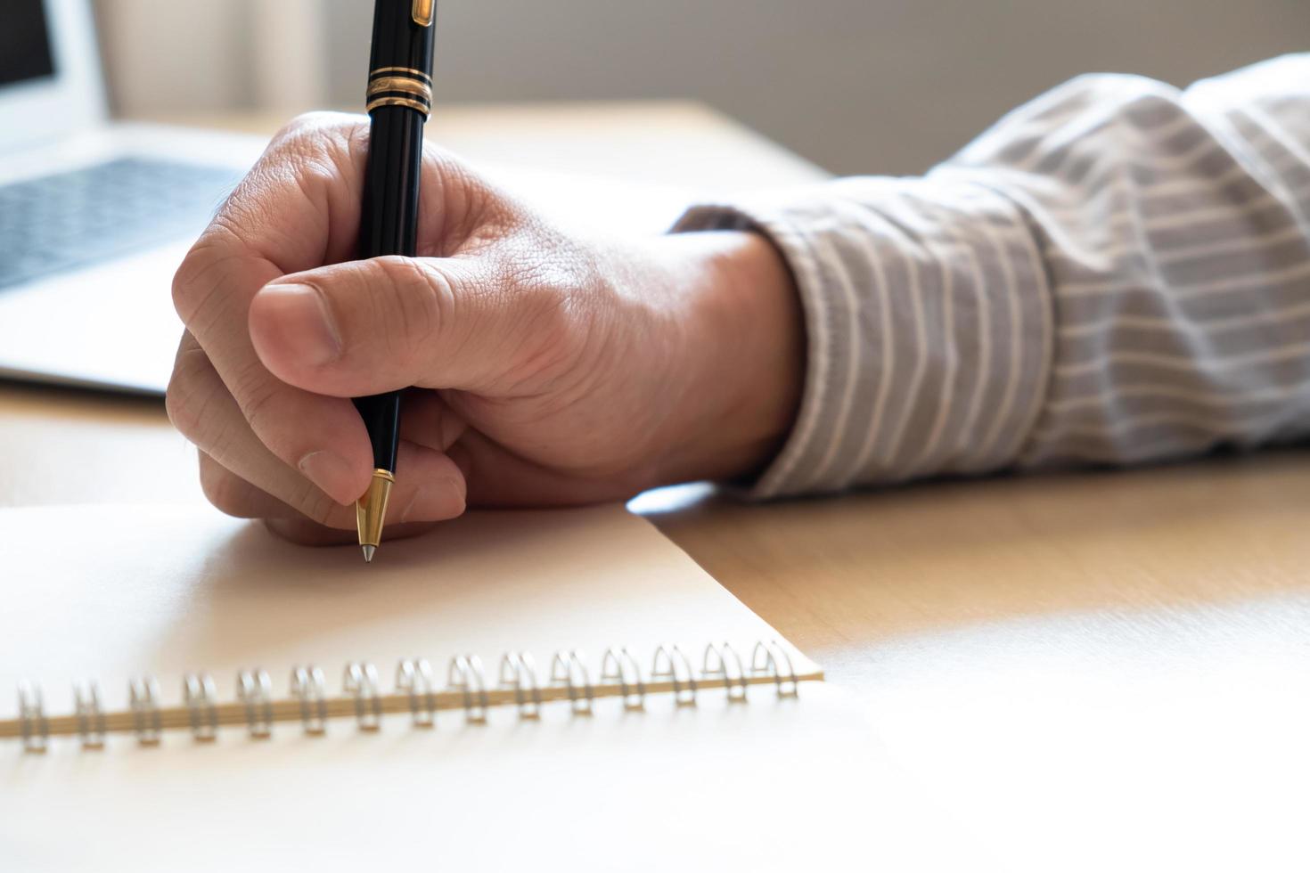 Selektiver Fokus auf die menschliche Hand mit Stift, um eine kurze Notiz auf Papier auf dem Arbeitstisch zu schreiben. mit verschwommenem Laptop-Computer im Hintergrund. Liste für die Planung oder Organisation von Aktivitäten während der Arbeitstage. foto