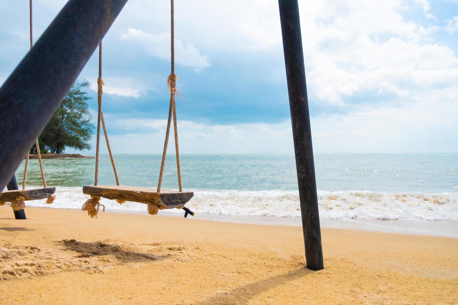 Selektiver Fokus auf Holzschaukel auf Sand am Strand. mit Wellen- und Meerblick mit Wolke, blauer Himmel. sommerferien auf der wunderschönen tropischen insel. Reiseziel. foto