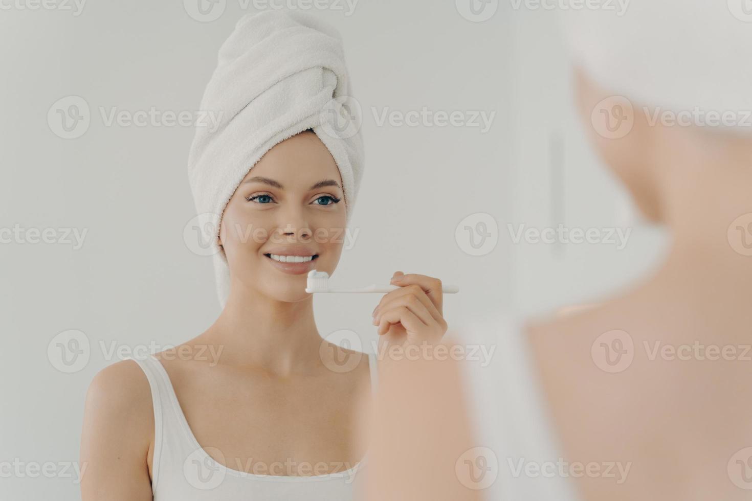 Gesunde schöne Frau mit Handtuch auf dem Kopf nach dem Duschen, die ein zahniges Lächeln hat, während sie sich sanft die Zähne putzt foto
