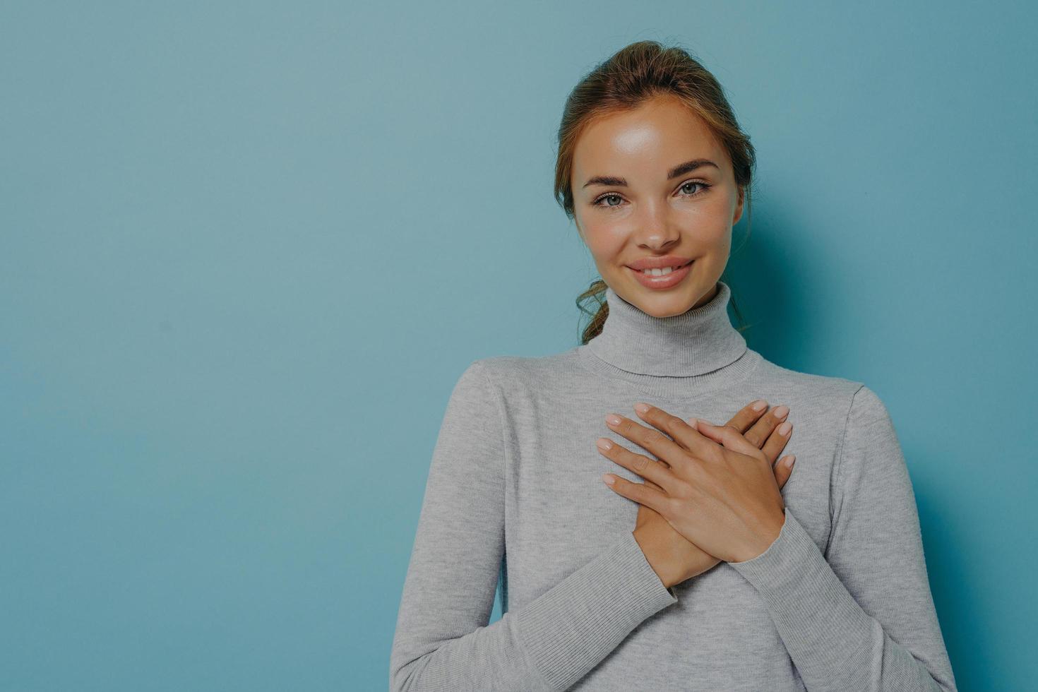 Attraktive junge lächelnde Frau, die gefaltete Hände in der Nähe des Herzens auf der Brust hält, während sie im Studio posiert foto