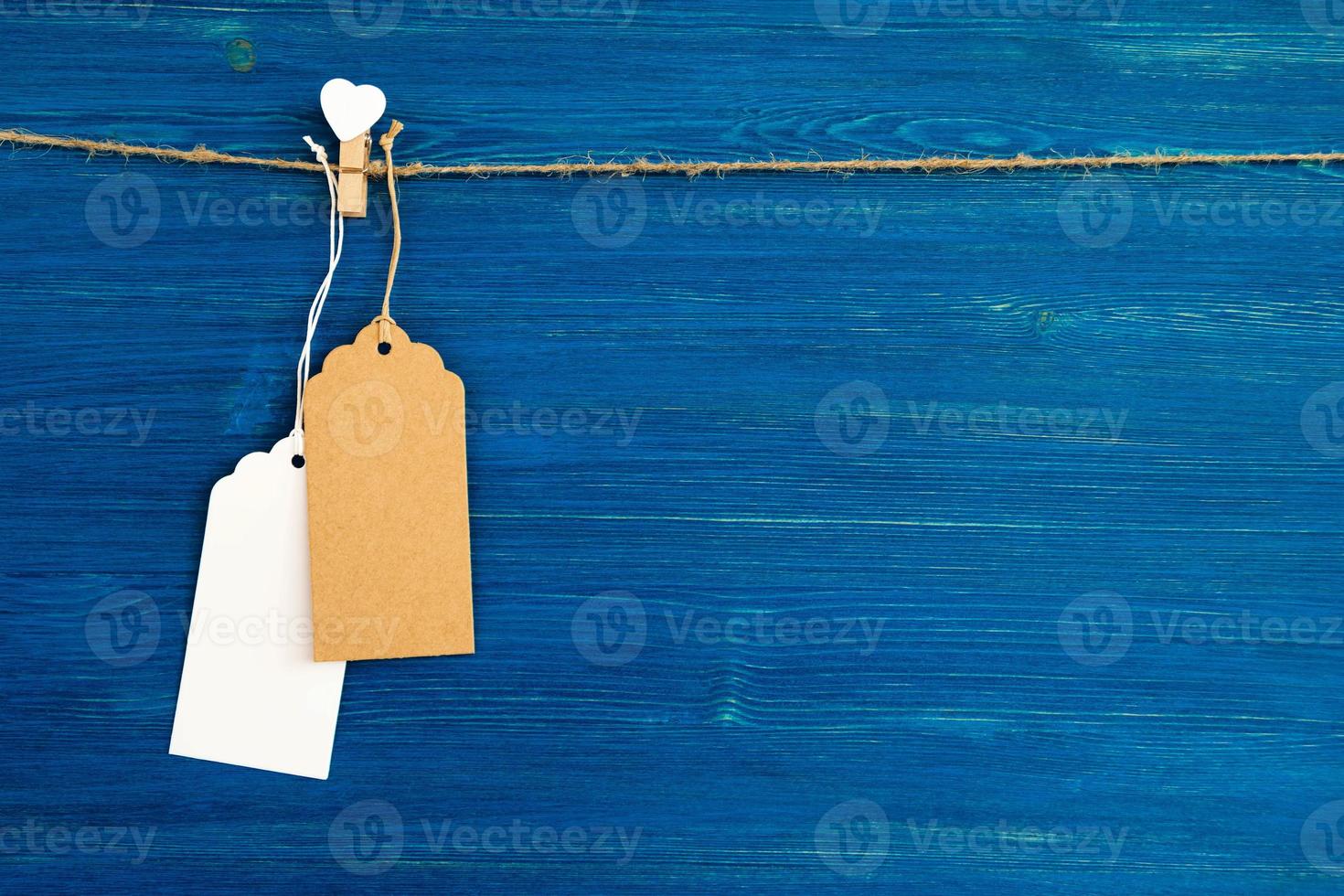 braune und weiße leere papierpreisschilder oder etiketten eingestellt und holzstift verziert auf weißem herz, das an einem seil auf dem blauen hölzernen hintergrund hängt. foto