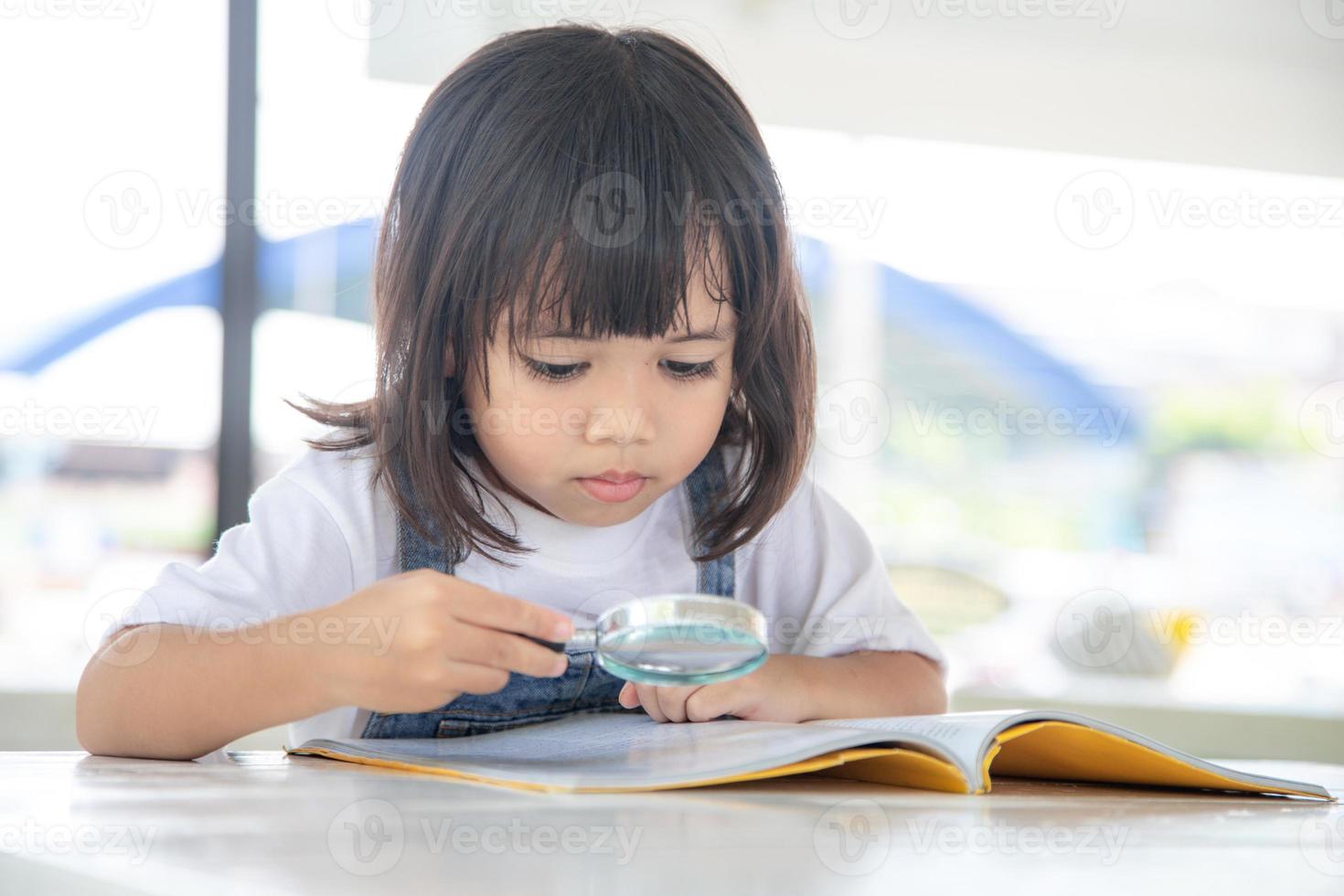 Asiatisches kleines Mädchen, das die Bücher auf dem Schreibtisch mit einer Lupe liest foto