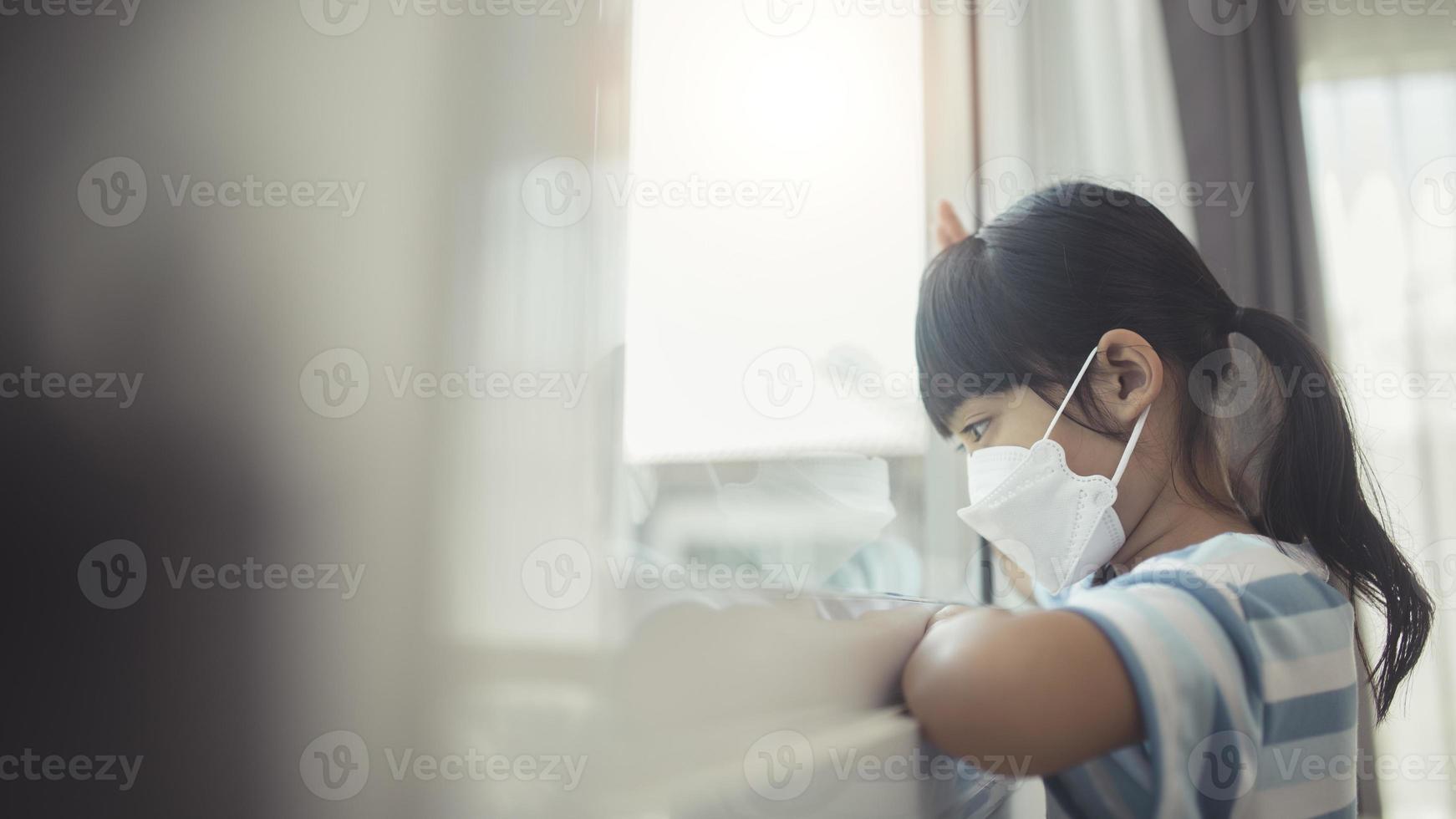 kleines asiatisches mädchen mit maske zum schutz von pm2.5 und covid-19. krankes kind kleine mädchen schauen mit sehnsucht auf die straße auf das fenster. Platz kopieren. Traurigkeitskind zu Hause isoliert foto