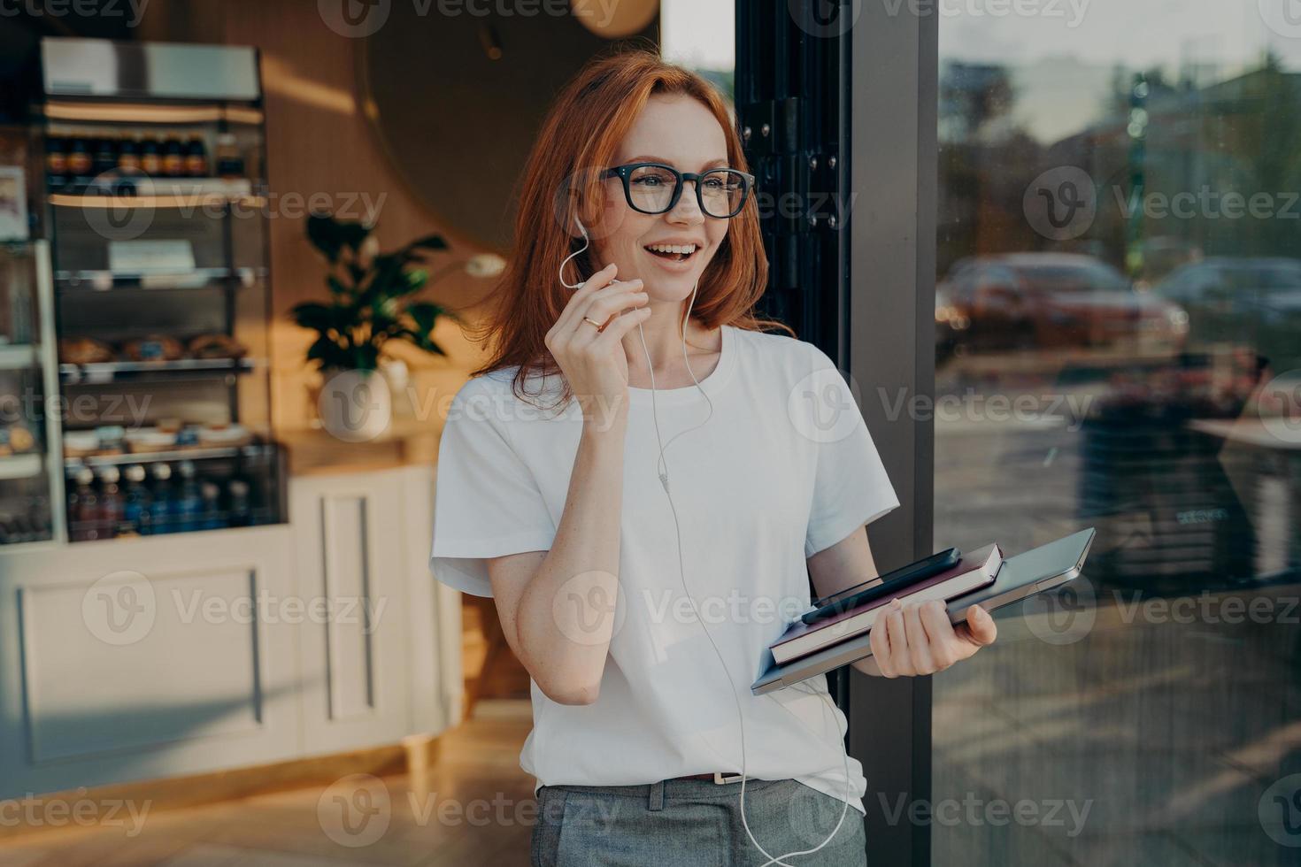 Fröhliche rothaarige Geschäftsfrau mit Laptop in der Hand, die auf dem Handy anruft und das Café verlässt foto