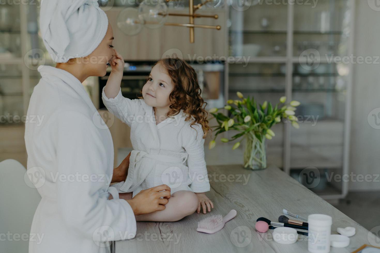 hübsches lockiges kleines mädchen berührt die nase ihrer mutter, die in einem weißen, weichen bademantel gekleidet ist, sitzt auf dem tisch mit kosmetischen produkten, die make-up für die mutter machen werden, die zusammen gegen ein gemütliches wohninterieur posiert foto