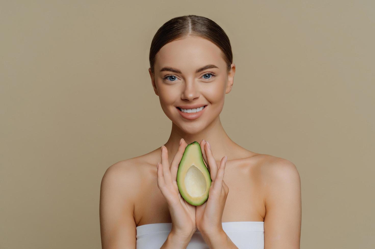 Schönheits- und Körperpflegekonzept. lächelnde ruhige europäerin mit gekämmten haaren hält avocado als empfehlung für eine nährende gesichtsmaske, die in ein badetuch gewickelt ist, isoliert über beigem studiohintergrund foto