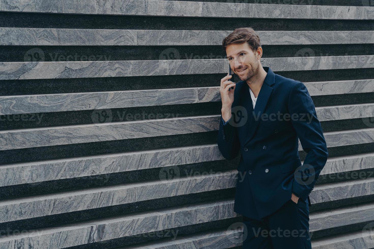 männlicher korporativer arbeiter im formellen schwarzen anzug hält die hand in der tasche und führt ein telefongespräch foto