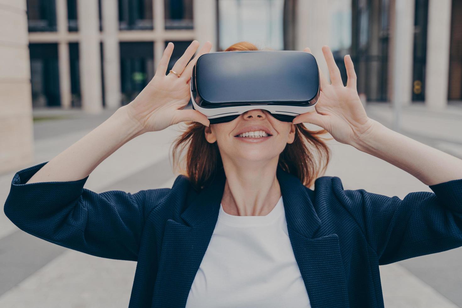 junge lächelnde rothaarige unternehmerin, die tragbare vr-brillen auf der stadtstraße testet foto