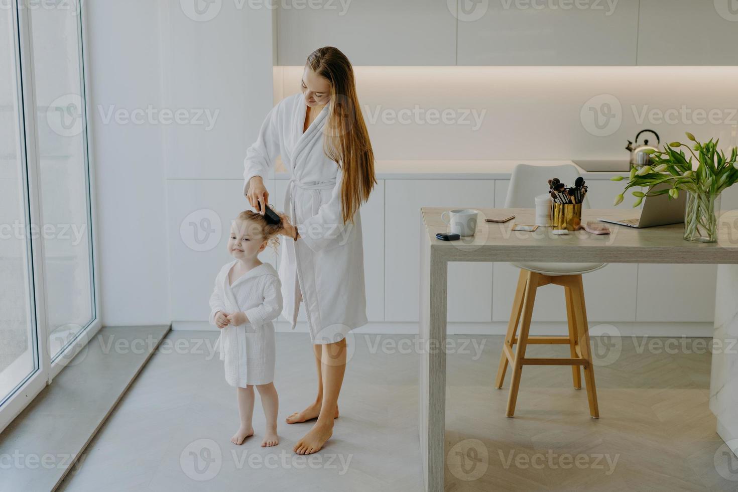 Die fürsorgliche Mutter kämmt die Haare ihrer kleinen Tochter und trägt weiße Bademäntel. schönes kleines lockiges Mädchen nach dem Duschen. Mutter macht dem Kind eine Frisur foto