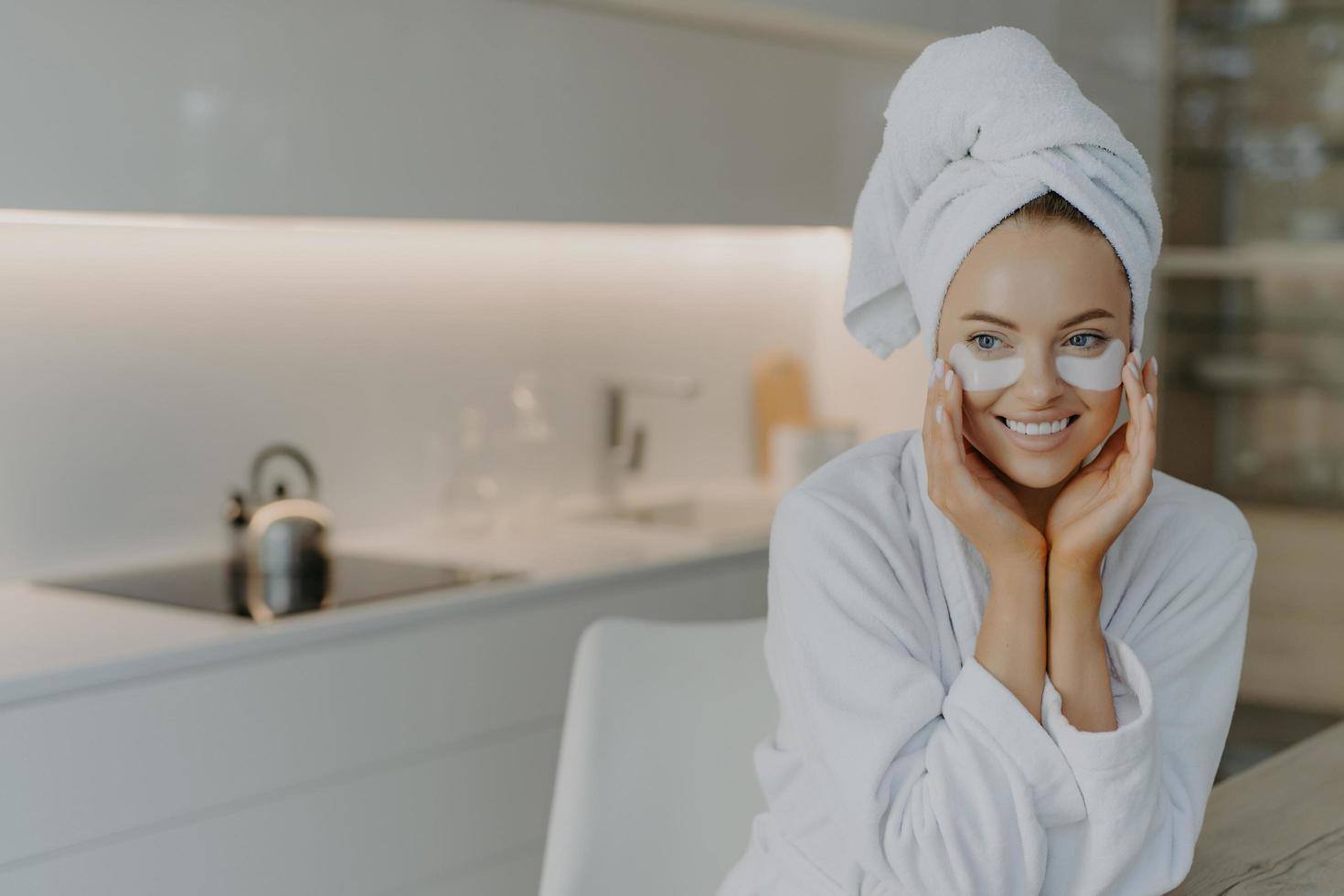 Foto einer schönen Frau mit gesunder Haut trägt Erholungspads unter den Augen auf, genießt die Hautpflege und eine wirksame Schönheitsbehandlung, gekleidet in einen weichen Bademantel, trägt ein eingewickeltes Handtuch auf dem Kopf und posiert über dem Innenraum