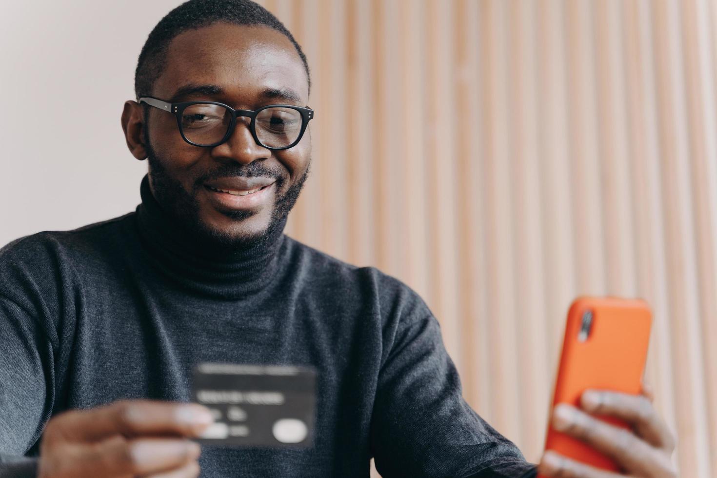 junger positiver afrikanischer ethnischer mannunternehmer in brille, der online mit kreditkarte bezahlt foto