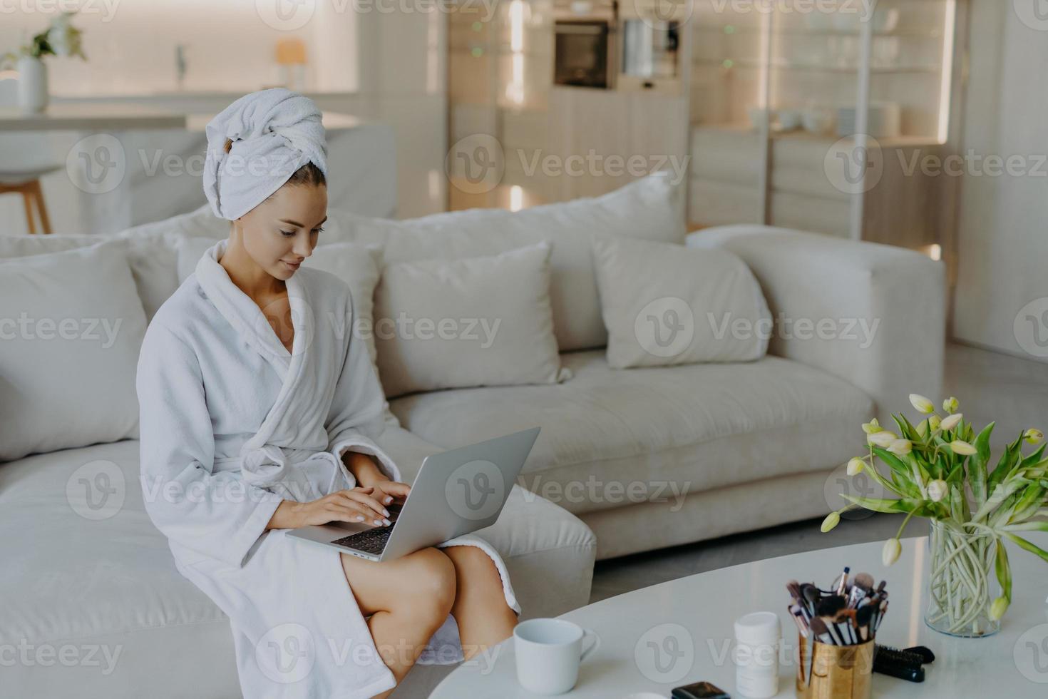 Foto einer schönen jungen Frau im Bademantel auf einem Laptop-Computer arbeitet weit entfernt von zu Hause, posiert auf einer bequemen Couch vor dem modernen Apartment-Interieur und trinkt Tee. Menschen Wohlbefinden Lebensstil