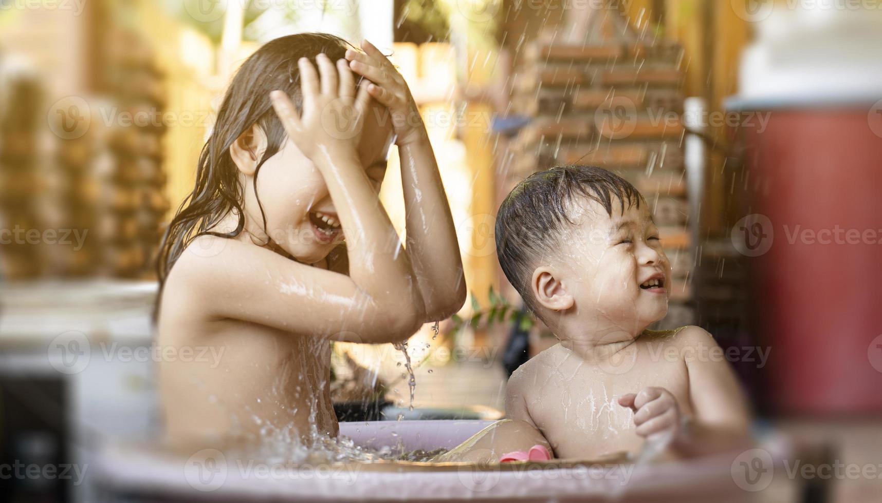 die ältere schwester und der bruder nahmen morgens glücklich eine badewanne aus plastik. Duschen Sie an einem heißen Tag draußen. Geschwister spielen gerne im Wasser. Kinder haben Spaß beim Spielen im Wasser foto