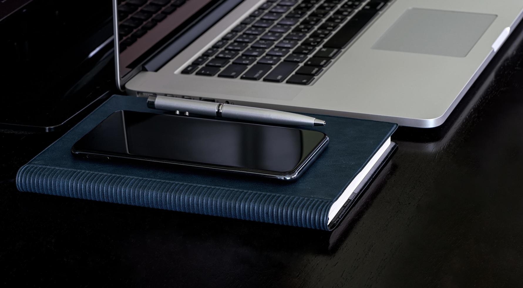 Laptop mit Smartphone und Notebook auf einem schwarzen Schreibtisch foto