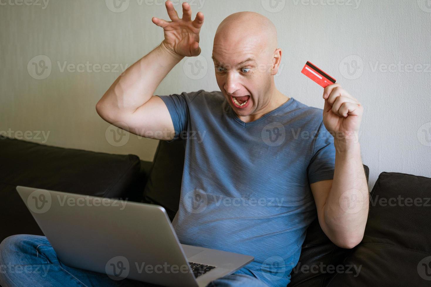 Emotionaler Glatzkopf auf Sofa mit Laptop und Kreditkarte in der Hand foto