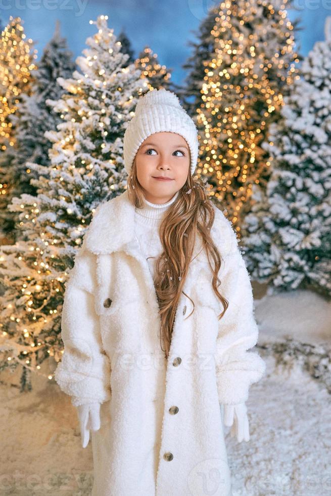 Süßes kaukasisches blondes Mädchen in weißem Öko-Pelzmantel, Hut und Handschuhen, das im Winterweihnachtswald spazieren geht. neues jahr, märchen, modekonzept foto