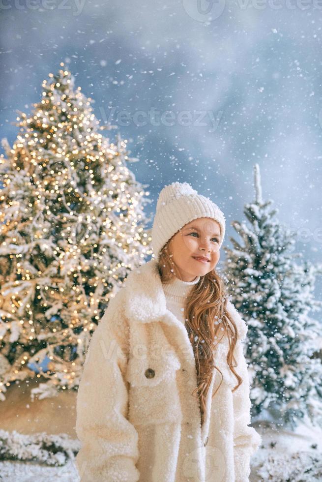 Süßes kaukasisches blondes Mädchen in weißem Öko-Pelzmantel, Hut und Handschuhen, das im Winterweihnachtswald spazieren geht. neues jahr, märchen, modekonzept foto