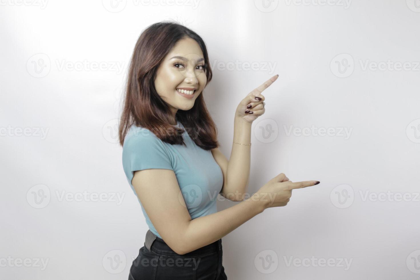 Aufgeregte asiatische Frau mit blauem T-Shirt, die auf den Kopierbereich neben ihr zeigt, isoliert durch weißen Hintergrund foto