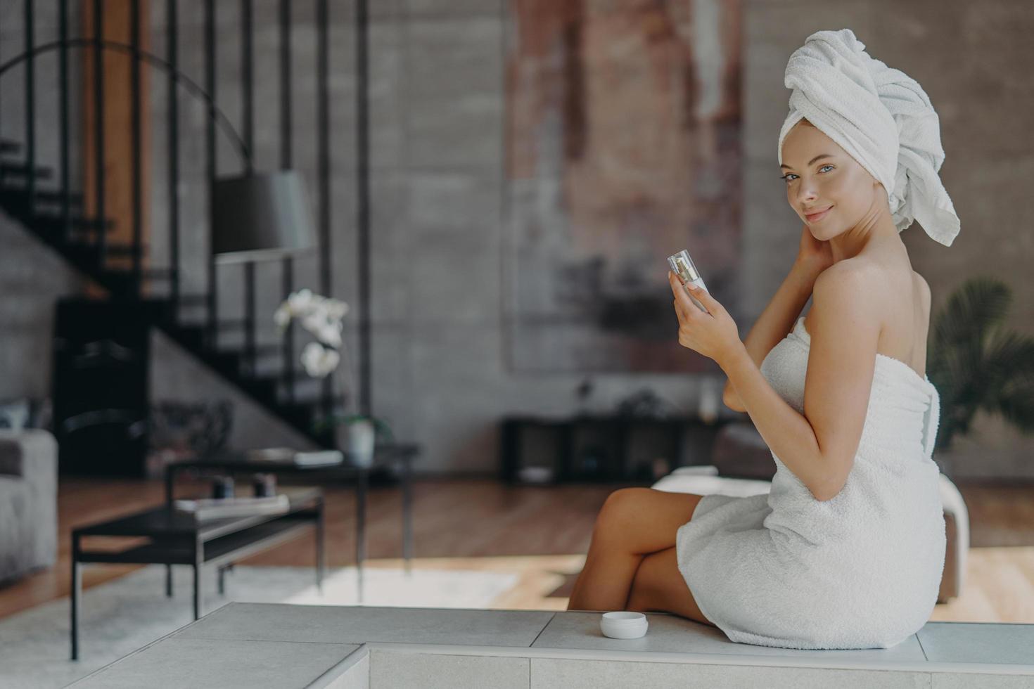 innenaufnahme einer gutaussehenden frau hält eine flasche lotion in der hand, hat gut gepflegte glatte haut trägt ein handtuch auf dem kopf und um den nackten körper nach dem duschen, posiert vor dem wohnraum. Beauty-Konzept. foto