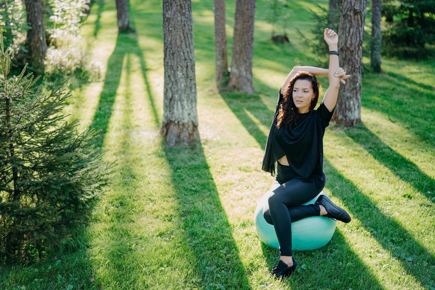 Foto einer aktiven, flexiblen, brünetten Frau, die sich mit Fitball dehnt, Pilates oder Fitnessübungen macht, an sonnigen Frühlingstagen im Wald auf grünem Gras posiert, an der frischen Luft gymnastische Übungen macht.