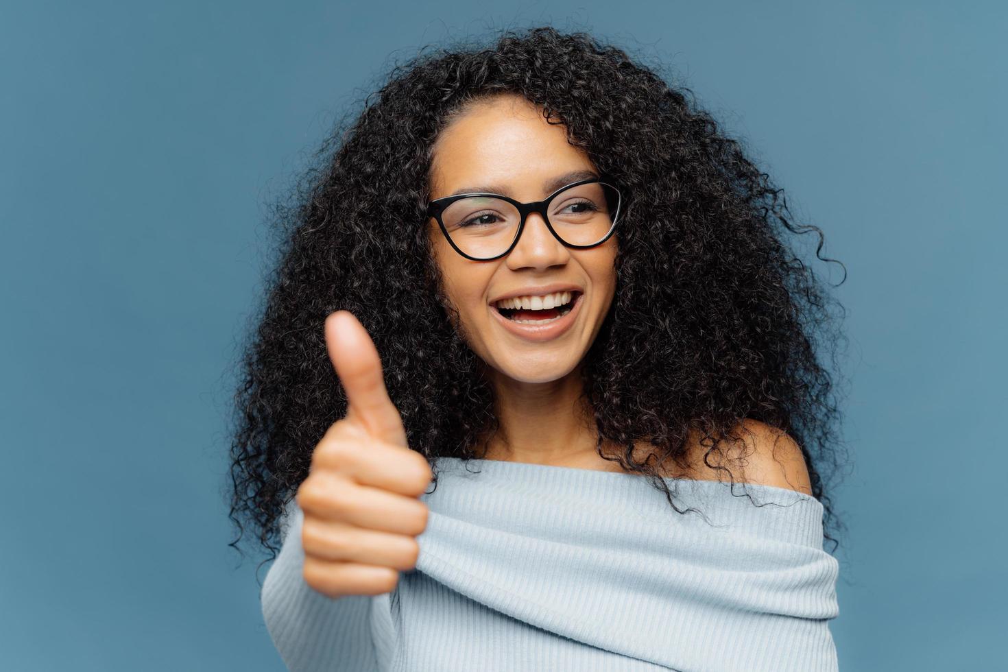 porträt einer glücklichen frau mit afrofrisur, zeigt daumen nach oben, gibt eine positive meinung ab, schaut zur seite, trägt eine optische brille und einen pullover, isoliert auf blauem hintergrund. Menschen, Vereinbarung, Zustimmung foto