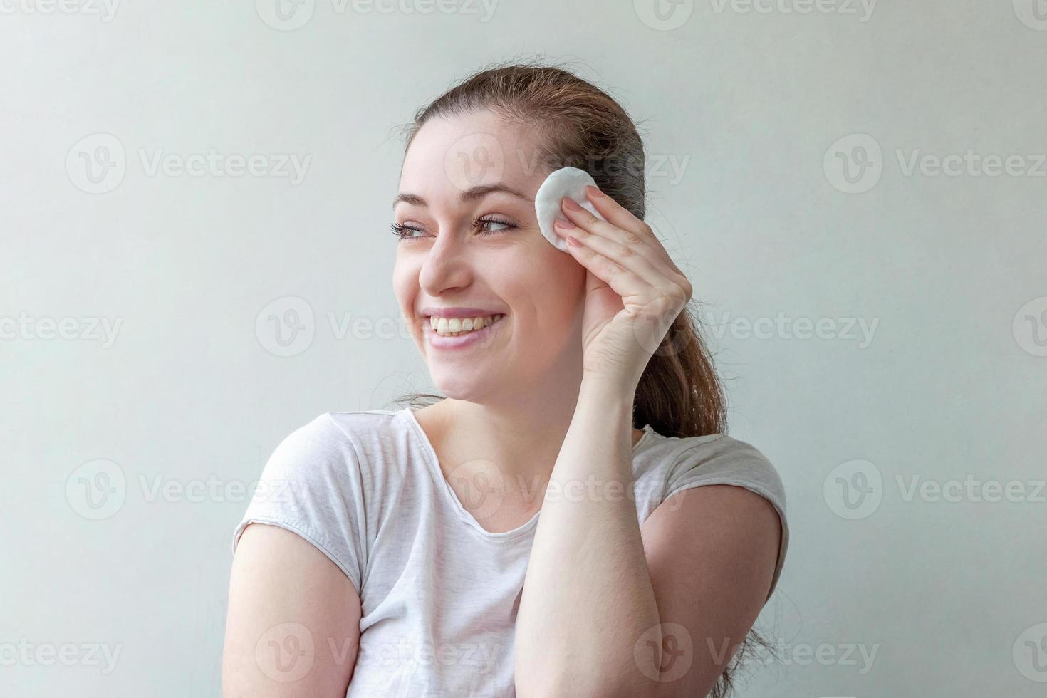 lächelnde Frau mit weicher, gesunder Haut, die Make-up mit einem Wattepad entfernt, das auf weißem Hintergrund isoliert ist foto