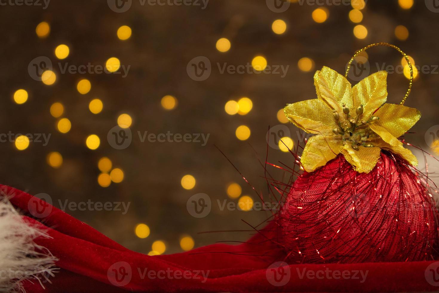 roter weihnachtsball mit goldener blume und roter weihnachtsmannmütze mit neujahrslichtern auf der rückseite. unscharfer Hintergrund. Urlaubspostkarte foto