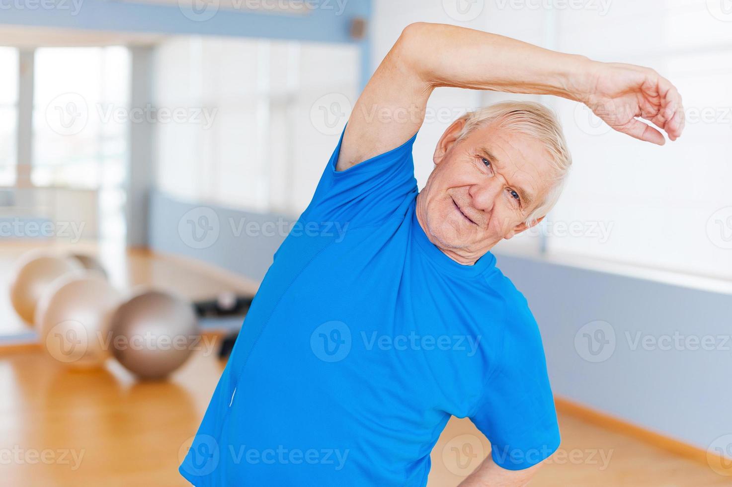 aktiver Senior. Fröhlicher älterer Mann, der Dehnübungen macht und lächelt, während er drinnen steht foto