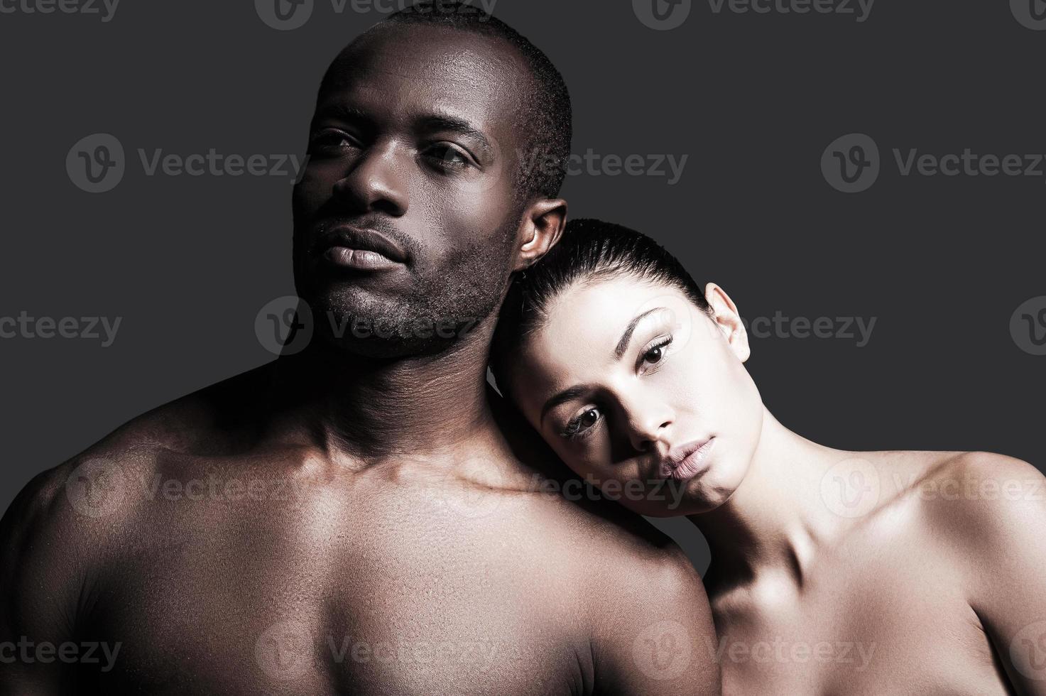 verschiedene Rassen und eine Liebe. Porträt eines hemdlosen afrikanischen Mannes und einer kaukasischen Frau, die sich aneinander binden, während sie vor grauem Hintergrund stehen foto