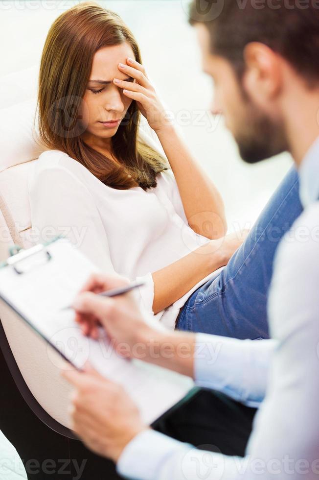 sie braucht einen fachkundigen rat. Depressive junge Frau, die auf dem Stuhl sitzt und die Hand auf dem Kopf hält, während der junge Mann neben ihr sitzt und etwas in seine Zwischenablage schreibt foto