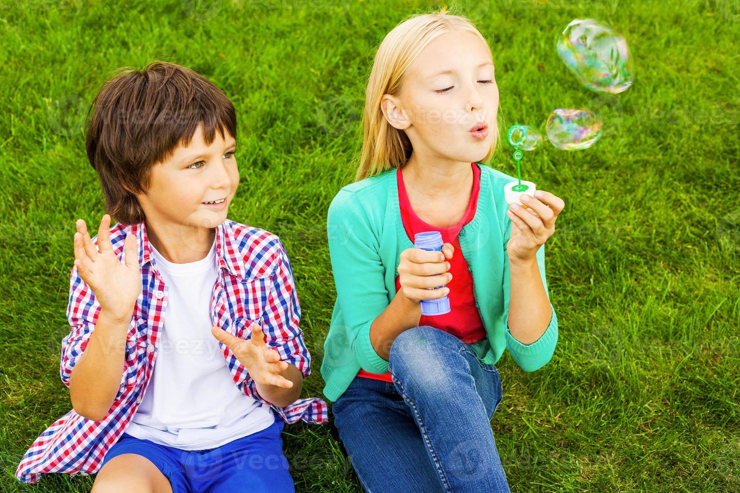 Blasenspaß. Zwei süße kleine Kinder, die Seifenblasen blasen, während sie zusammen auf dem grünen Gras sitzen foto