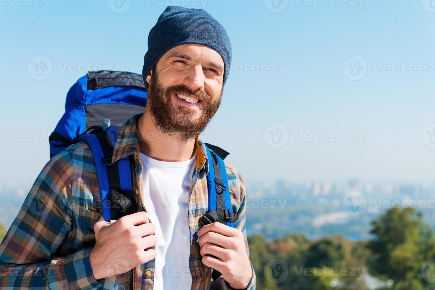 glücklicher Tourist. hübscher junger Mann mit Rucksack und Blick in die Kamera mit einem Lächeln, während er in der Natur steht foto