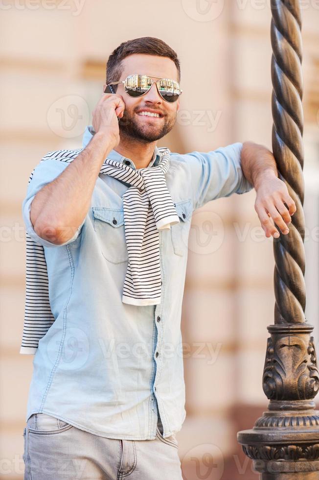 nettes Gespräch. hübscher junger Mann mit Sonnenbrille, der auf dem Handy spricht und lächelt, während er sich im Freien an die Säule lehnt foto