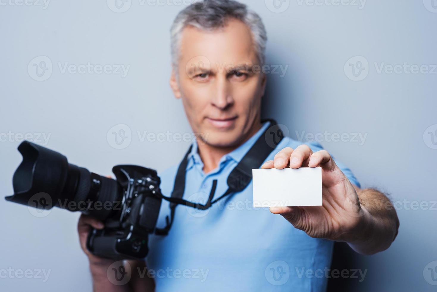 professioneller Fotograf. Porträt eines selbstbewussten, reifen Mannes im T-Shirt, der eine Kamera hält und die Visitenkarte ausstreckt, während er vor grauem Hintergrund steht foto