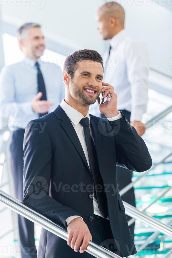 Tolle Neuigkeiten aus einem niedrigen Winkel eines selbstbewussten jungen Mannes in Abendkleidung, der am Handy spricht und lächelt, während er sich mit Menschen im Hintergrund die Treppe hinunterbewegt foto
