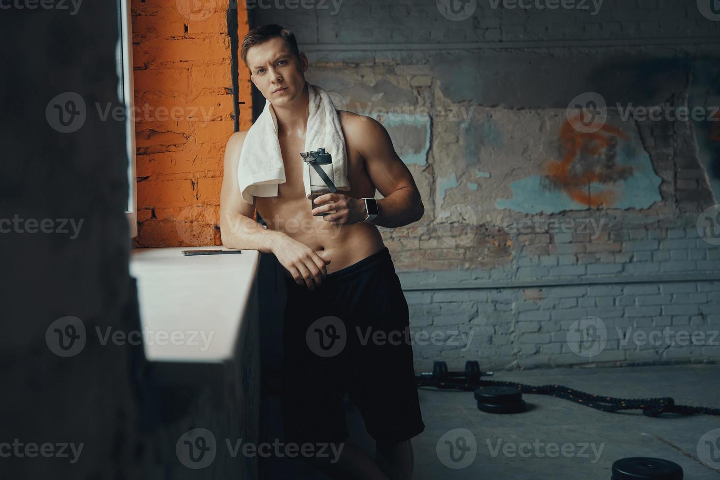 Selbstbewusster muskulöser Mann, der eine Flasche mit Wasser hält, während er in der Nähe des Fensters im Fitnessstudio steht foto