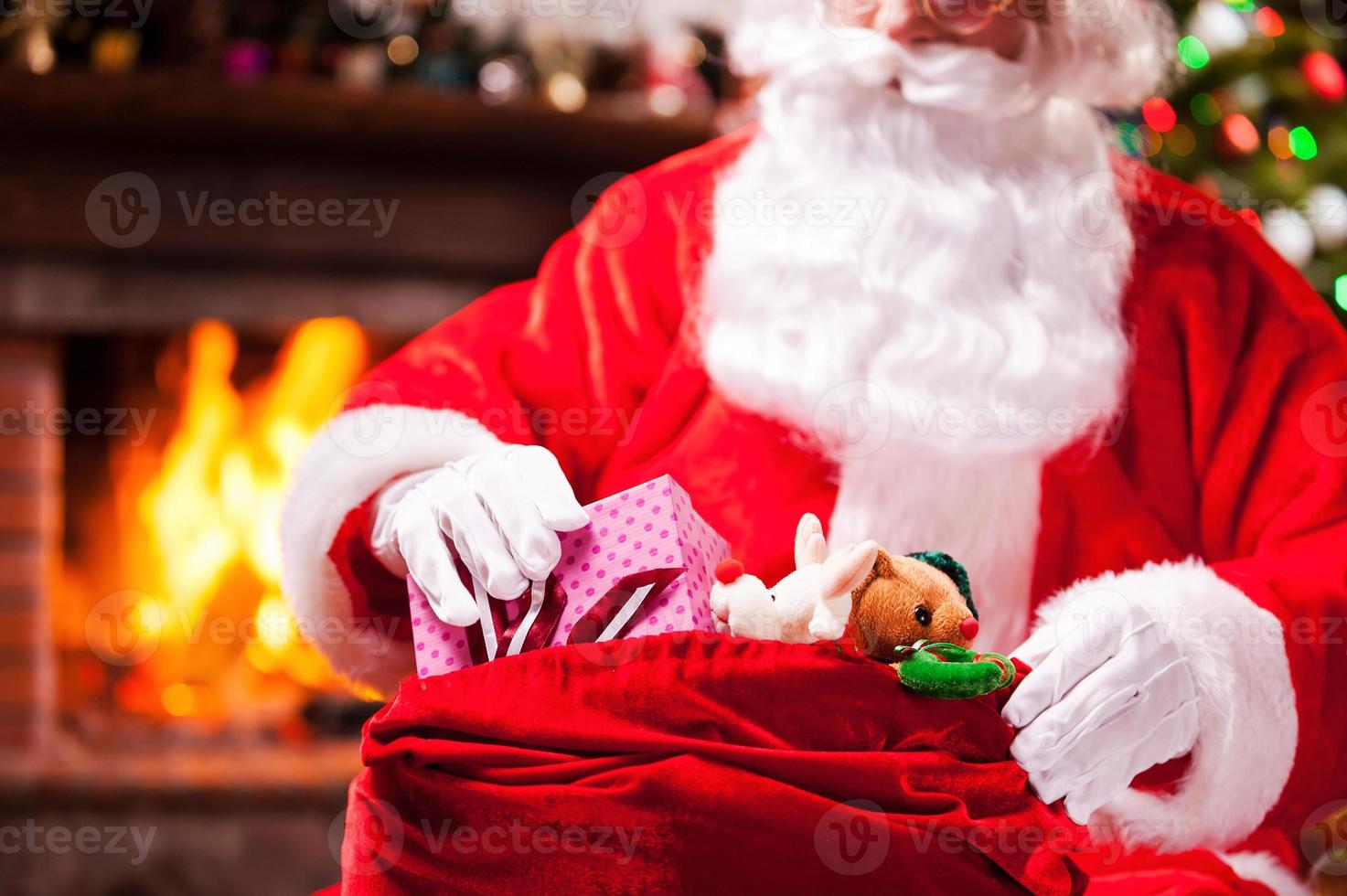 weihnachtsgeschenke verpacken. Nahaufnahme des Weihnachtsmanns, der Weihnachtsgeschenke in den Sack packt, mit Kamin und Weihnachtsbaum im Hintergrund foto