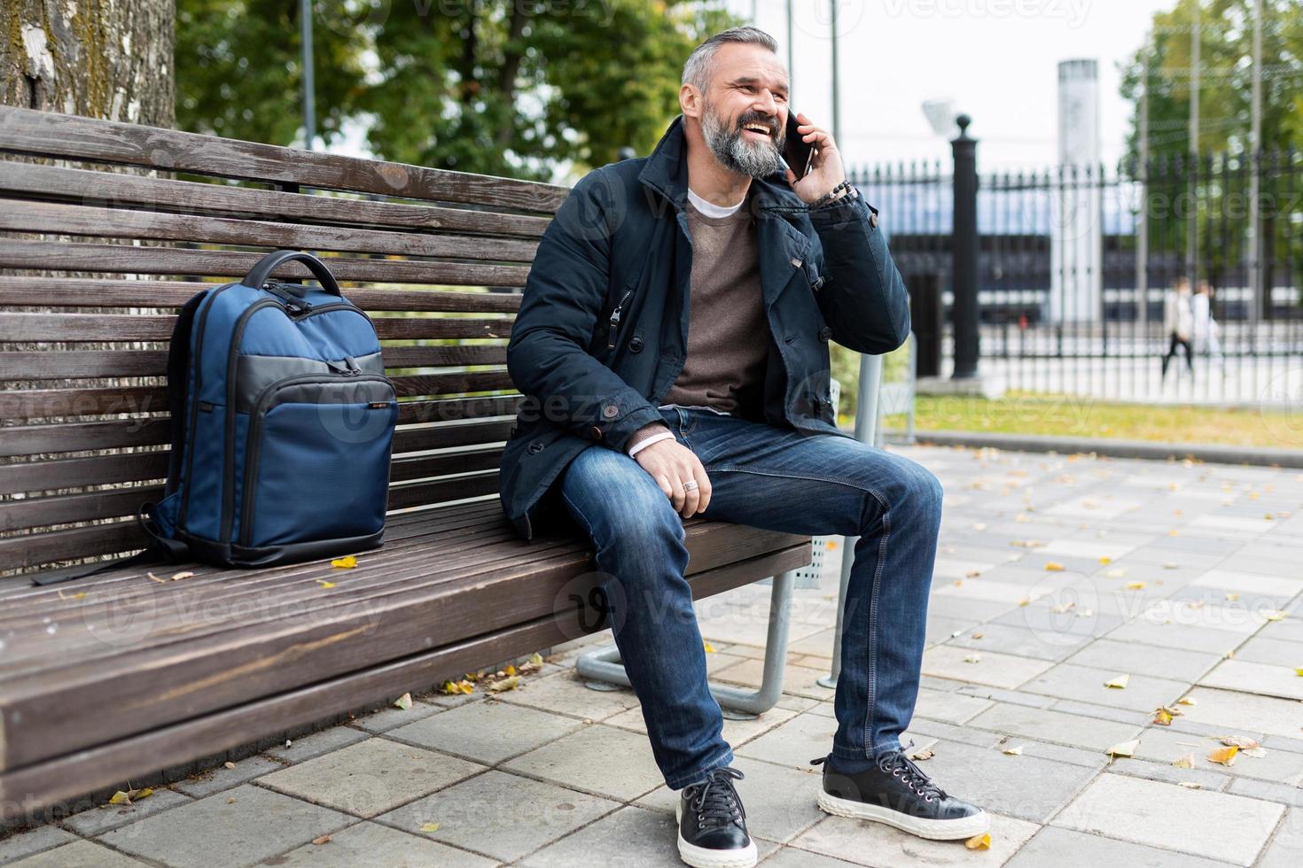 Ein Mann mit grauem Bart telefoniert, während er im Park auf einer Bank sitzt foto