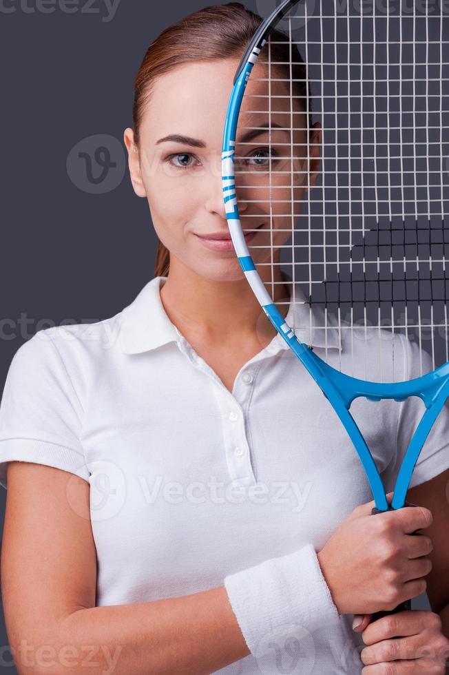 Ich bin im Spiel. Schöne junge Frauen in Sportkleidung, die einen Tennisschläger vor die Hälfte ihres Gesichts halten und die Kamera betrachten, während sie vor grauem Hintergrund stehen foto