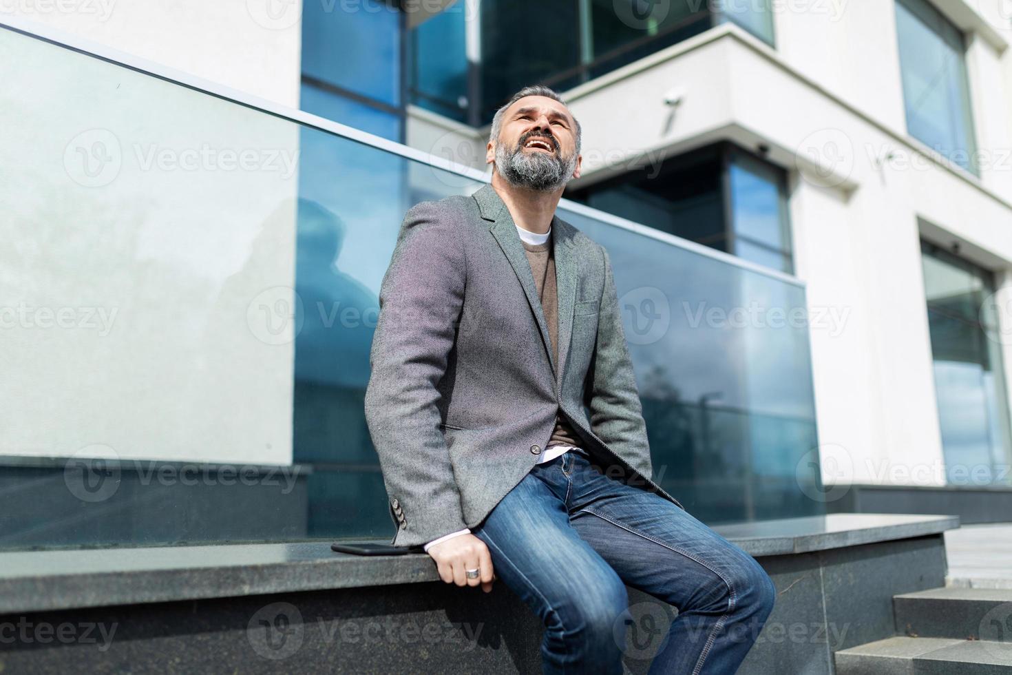 Älterer Geschäftsmann mit grauem Bart sitzt in der Nähe des Bürogebäudes und schaut nach oben, Konzept des Geschäftserfolgs und des Karrierewachstums foto