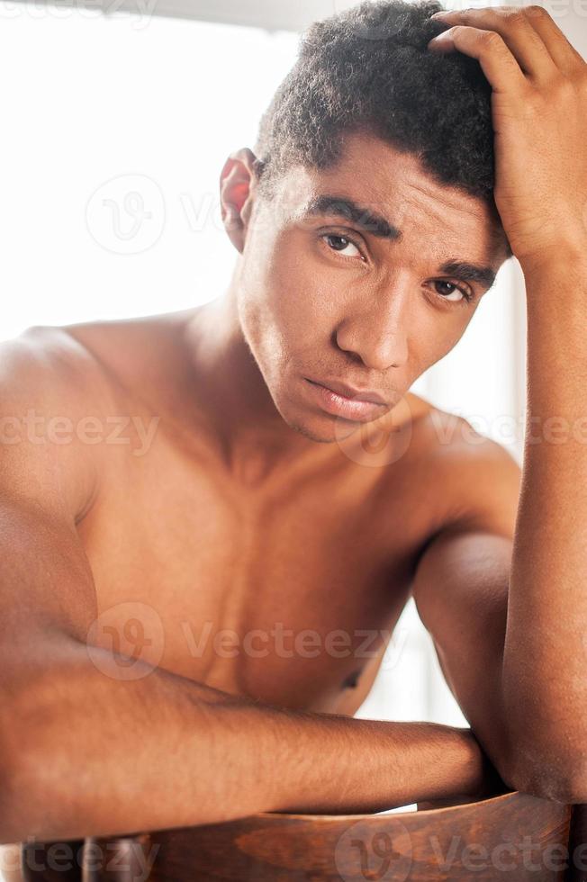 durchdringender Blick. hübscher junger afroamerikanischer mann, der auf dem stuhl sitzt und in die kamera schaut, die hand auf seinem kopf hält foto