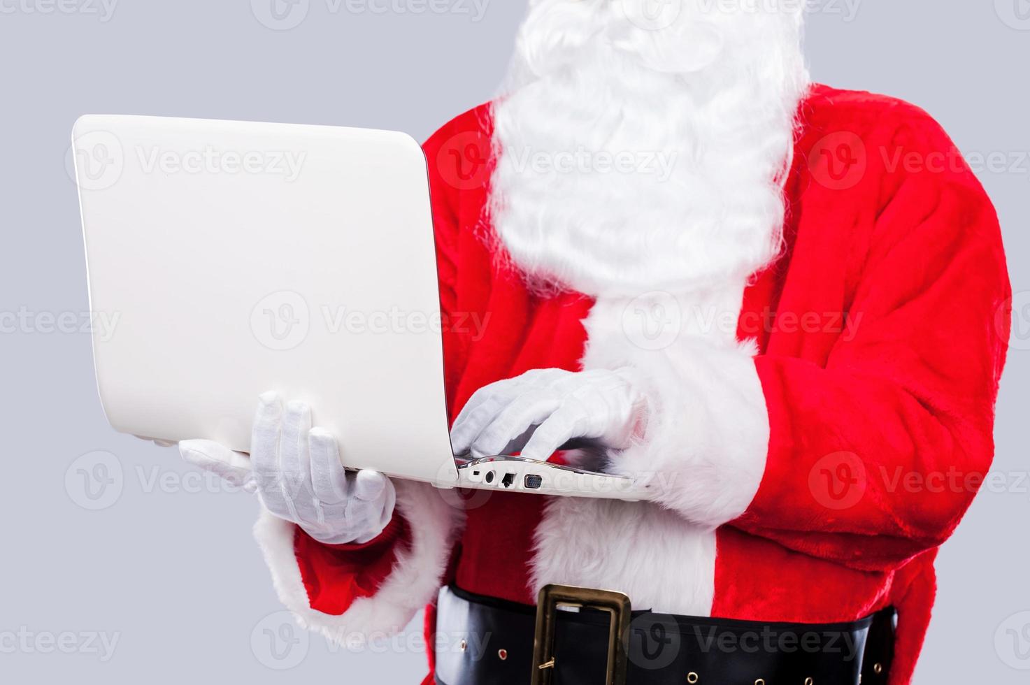 digitaler zeitalter weihnachtsmann. weihnachtsmann, der am laptop arbeitet, während er vor grauem hintergrund steht foto